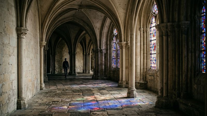 Sunlit stone ambulatory with slender columns and coloured glass wash, suggesting a twelfth-century French abbey choir without naming a specific building.