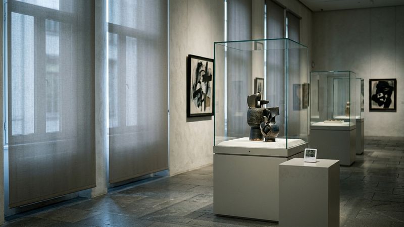 Dim museum gallery with lowered blinds, sealed display cases and a small climate sensor on a plinth, cool north light, no readable labels