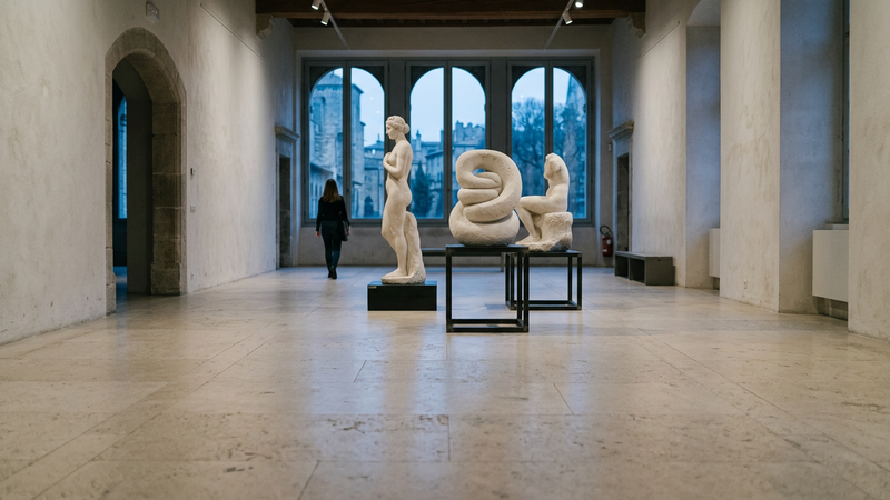 Sunlit museum gallery with overlapping marble sculptures and a wide empty stone floor, distant windows dissolving into pale blue haze, no text or logos