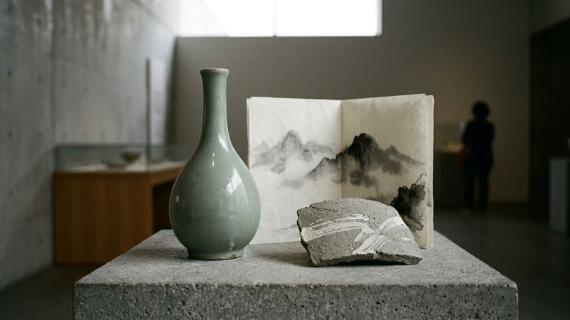 Grey-green celadon glaze on a curved ceramic shoulder beside white slip and ink-wash mountain tones in cool museum light