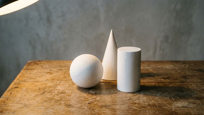 Studio still life of plaster sphere, cylinder and cone on a wooden table under a single directional lamp, with clear highlights and cast shadows, no text.