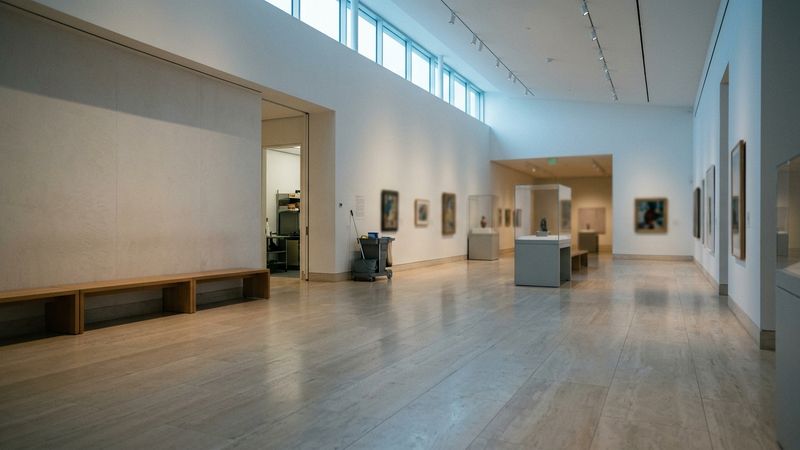 Sunlit museum gallery with empty benches and pale stone floors, collection displays soft in the distance, quiet before opening