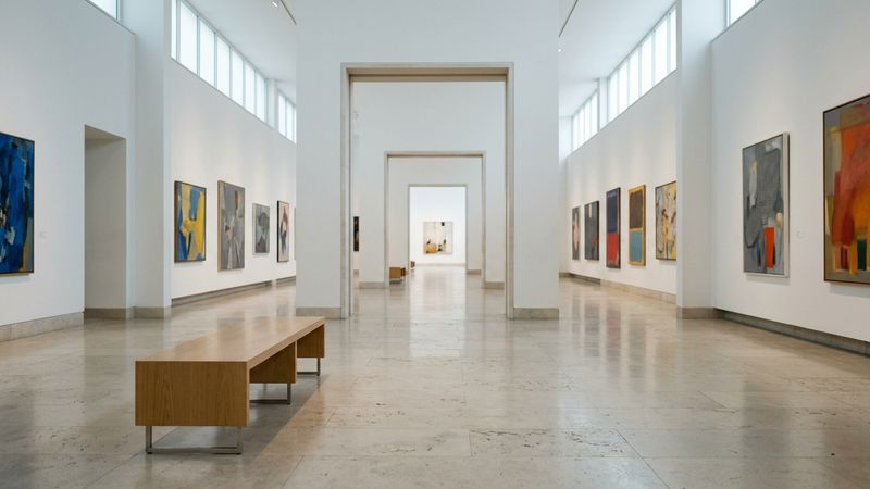 Sunlit museum gallery with empty benches, pale walls and framed paintings along a quiet enfilade, no visitors in frame
