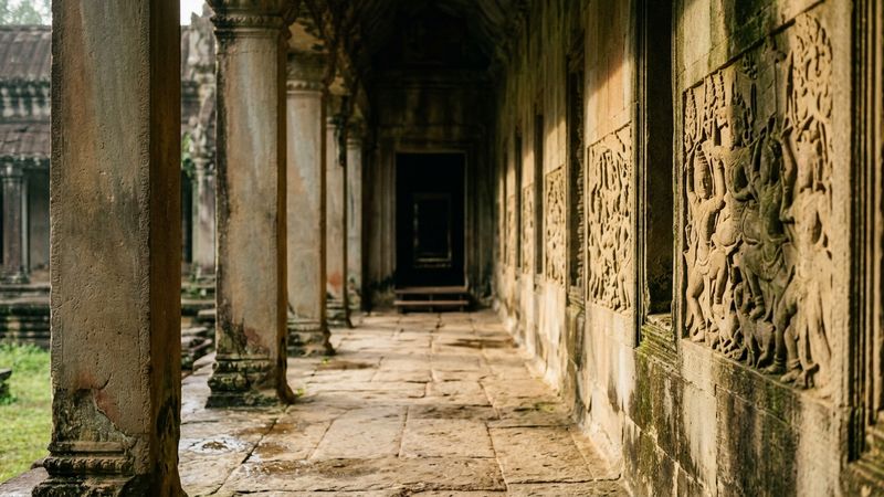 Weathered sandstone temple gallery in warm monsoon light, carved narrative reliefs along a shadowed corridor, no readable inscriptions